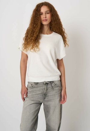 Junge Frau mit langen lockigen Haaren, die ein lockeres weißes kurzärmliges Oberteil und hellgraue hoch taillierte Jeans vor einem schlichten Hintergrund trägt.