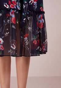 Falda negra translúcida con un patrón floral en rojo, blanco y azul sobre piernas desnudas contra un fondo beige claro.