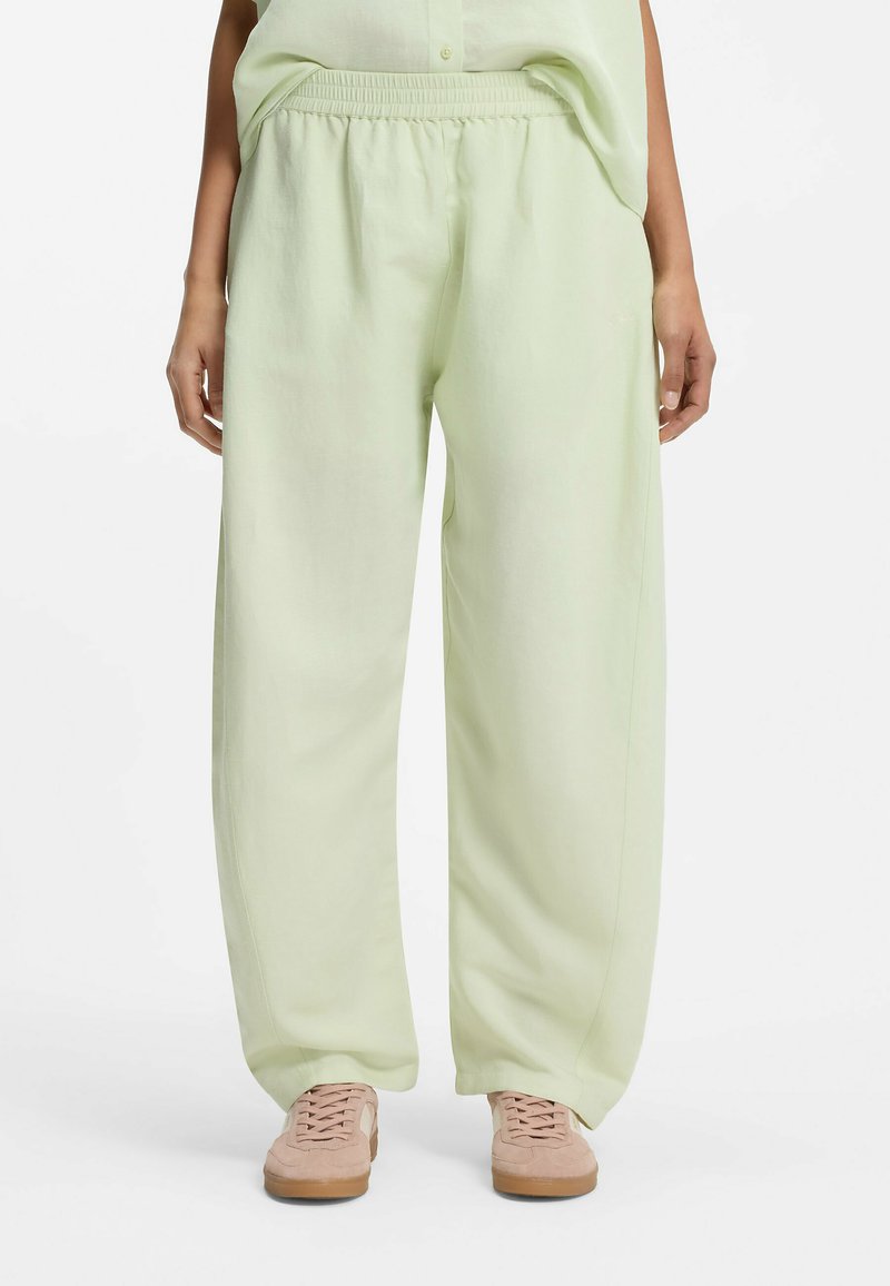 Personne portant un pantalon à taille élastique vert clair, coupe ample, associé à des baskets rose pâle, debout contre un fond blanc.