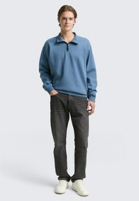 Hellblaues, langärmliges Pullover mit Reißverschlusskragen, kombiniert mit dunkelgrauen Jeans und weißen Sneakers. Lässige Passform, weicher Stoff.