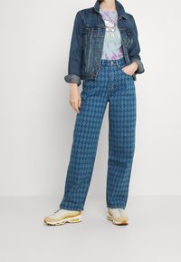 Kobieta w dżinsowej kurtce, koszulce z tie-dye z motywem Hello Kitty, wzorzystych szerokich dżinsach i białych oraz żółtych sneakersach, stojąca z jedną ręką w kieszeni.