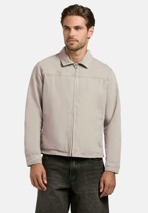 Mann iført lys beige glidelåsjakke med krage og mørkegrå bukser, stående mot en ensfarget hvit bakgrunn.