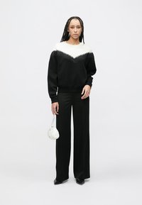 Črna jopa z belim, teksturiranim V-vzorcem na izrezu, kombinirana s črnimi hlačami širokih hlačnic in majhno belo torbico.