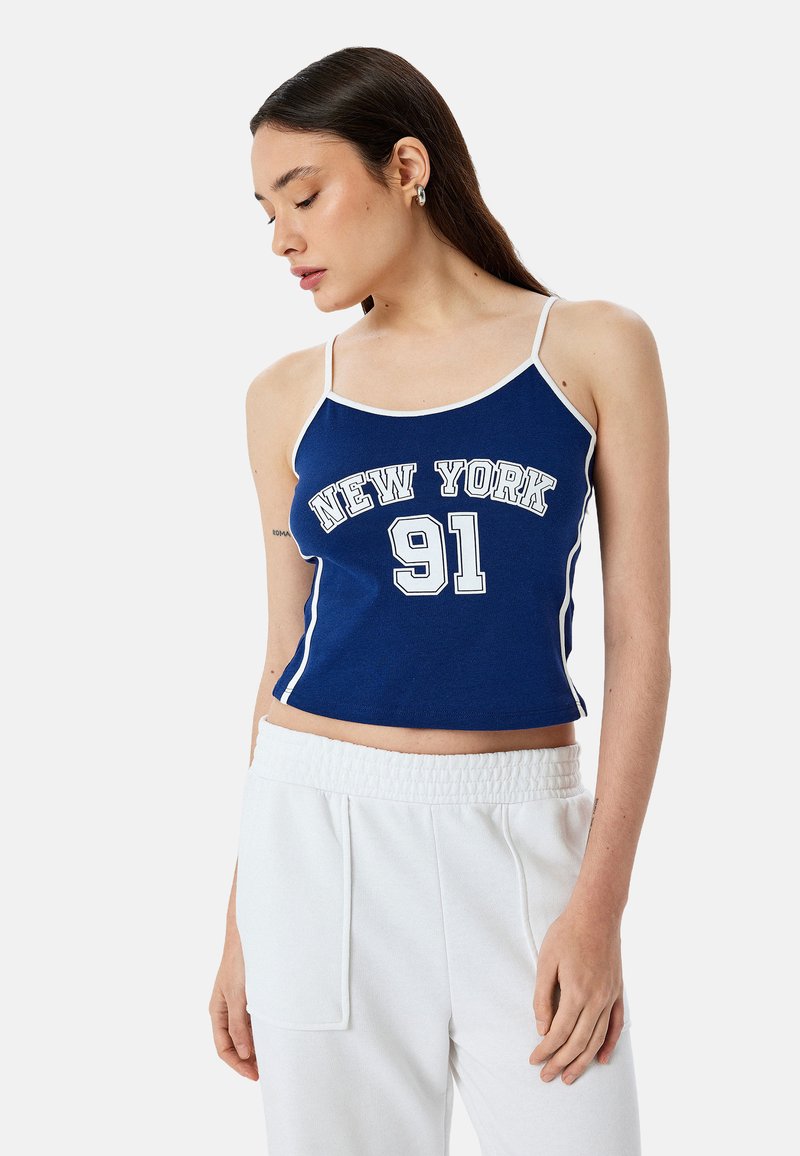 Blauwe crop top met witte afwerking waarop "NEW YORK" en "91" in vet afgerukte lettertype staan. Het materiaal lijkt zacht en goed aansluitend.