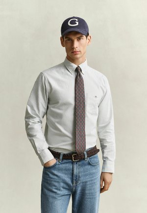 Homme portant une chemise à rayures boutonnée, une cravate à motifs, un jean bleu, une ceinture tressée foncée et une casquette bleu marine avec la lettre G, debout avec une main dans la poche.
