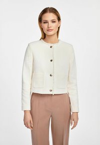 Biała, fakturowana krótka kurtka z okrągłym dekoltem, zapięcia na srebrne guziki i dwiema przednimi kieszeniami, zestawiona z jasnoróżowymi spodniami.