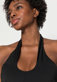 Top halter preto com um decote profundo, textura suave e costuras mínimas, exibindo um design elegante e ajustado.