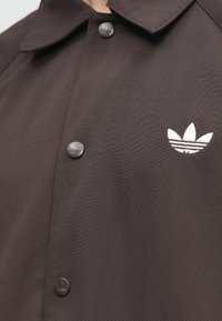 Καφέ σακάκι με κουμπιά και γιακά, με δύο σκούρα κουμπιά και λευκό κεντημένο λογότυπο στην αριστερή πλευρά.