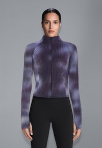 Femme portant un haut ajusté à manches longues avec fermeture éclair, motif tie-dye violet et gris, et un pantalon noir taille haute, sur un fond gris.