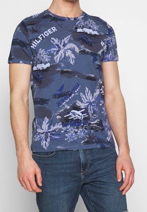 T-shirt bleu à manches courtes avec un imprimé tropical de feuilles de palmier, bateaux et texte de marque dans différentes nuances de bleu et de blanc.