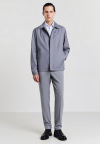 Giacca grigia con colletto, tasche frontali e vestibilità rilassata, abbinata a pantaloni grigi su misura e stivaletti neri alla caviglia. Tessuto dalla superficie liscia.