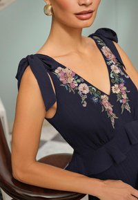 Rochie bleumarin cu broderie florală pe decolteu în formă de V, material texturat și bretele legate la umeri. Prezintă un tiv peplum.