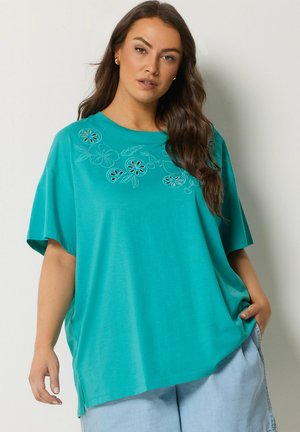 Femme aux longs cheveux bruns portant un t-shirt ample bleu sarcelle avec un motif floral découpé et un pantalon bleu clair, posant avec une main dans la poche.