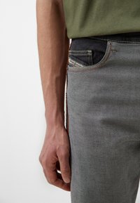 Vaqueros de mezclilla gris con una cinturilla oscura, acentos de costura roja y una etiqueta de bolsillo visible. Mano colocada a un lado para soporte.