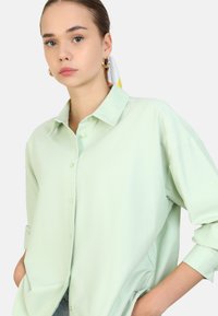 Camisa abotonada de color verde claro con un corte relajado, mangas largas y cuello. Presenta una tela suave y ligera con un sutil brillo.