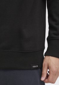 Sort sweatshirt med ribbede manchetter og kant. Har et lille sort mærke med hvid tekst. Glat tekstur, tætsiddende design.