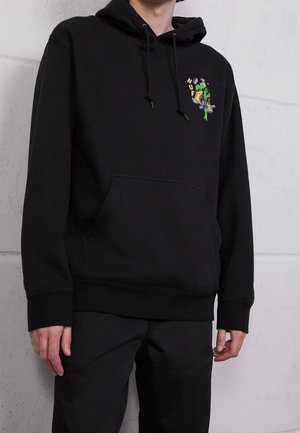 Person iført en sort hættetrøje med forlomme og en lille, farverig grafik på venstre bryst, stående mod en lys grå væg.