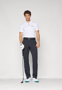 Hvit kortermet polo med glidelås og svart logo, sammen med mørke bukser. Golfhanske på venstre hånd, holder en golfkølle, iført sko med blå aksenter.