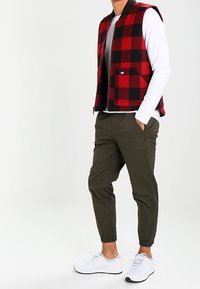 Man som har på sig en vit långärmad skjorta, väst i röd och svart rutig design, olivgröna joggerbyxor och vita sneakers, står med händerna i fickorna.