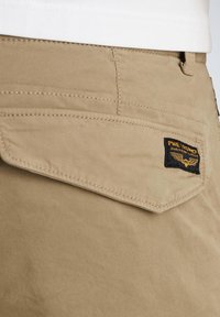 Beige katoenen shorts met een klepzak met een zwart label, zichtbare stiksels en een gladde textuur. Focus op functionele ontwerpelementen.