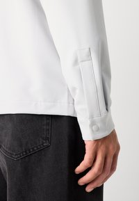 Cămașă albă cu mâneci lungi și mansete ajustate, cu închidere prin capse. Materialul pare neted, fără modele vizibile, asortată cu blugi închisi la culoare.