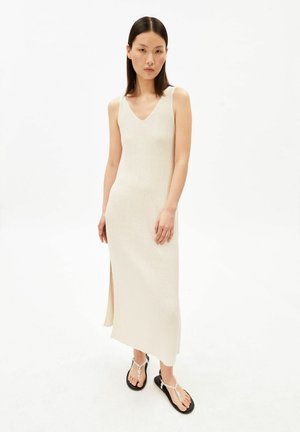 Junge Frau mit langen, glatten Haaren, die ein ärmelloses, geripptes, cremefarbenes Midikleid und schwarze Sandalen mit weißen geflochtenen Riemen trägt.