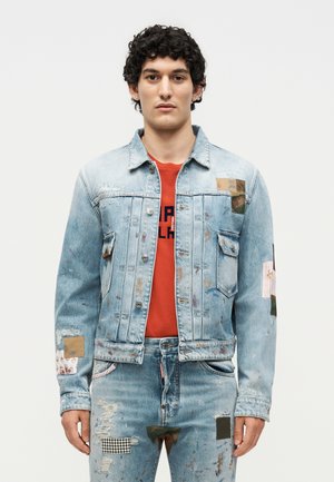 Άνδρας που φορά ανοιχτό μπλε τζιν σακάκι και τζιν με πιτσιλιές μπογιάς και πατσάκι, σε συνδυασμό με κόκκινη γραφιστική μπλούζα, στέκεται μπροστά σε λευκό φόντο.