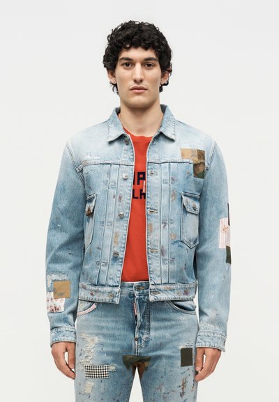 Man bär en ljusblå denimjacka och jeans med färgstänk och lappning, kombinerat med en röd grafisk t-shirt, står mot vit bakgrund.