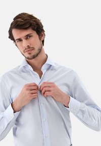 Hellblaues Hemd mit Spread-Kragen, glatter Textur und kontrastierenden weißen Knöpfen. Modell befestigt das Hemd durch Knöpfen.