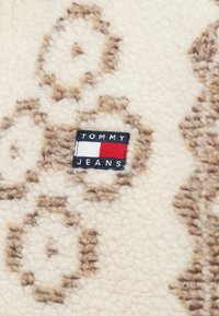 Kremkrāsas flīsa audums ar brūniem ziedu rakstiem un taisnstūra "TOMMY JEANS" etiķeti, kas atainota sarkano, balto un zilo krāsu toņos. Mīksta tekstūra.