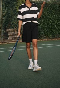Polo a righe bianche e nere, pantaloni corti neri, calze bianche e scarpe da ginnastica. Tiene una racchetta da tennis, su un campo verde.