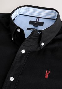 Črna bombažna srajca na knofe z belimi gumbi in rdečo vezenino jelena. Notranji ovratnik ima modro-bele črte.