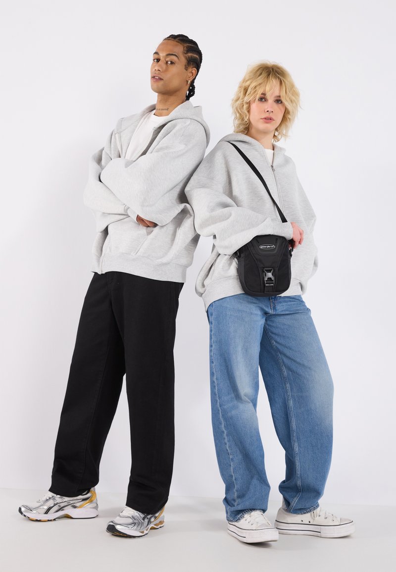 Sudaderas grises de gran tamaño, pantalones holgados negros y azules, y zapatillas deportivas. Un modelo sostiene un pequeño bolso cruzado negro. Fondo blanco liso.