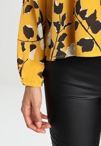 Blouse à motif floral jaune en tissu léger, avec des manches longues à poignets froncés, associée à un pantalon noir ajusté.