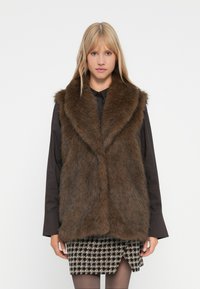 Vrouw in een bruine faux fur vest over een zwart langemouw shirt en een zwart-wit geblokte rok voor een witte achtergrond.