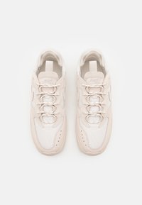 Beige athletic shoes with a textured mesh upper, padded collar, and light laces; features stitched accents and side logo detailing.