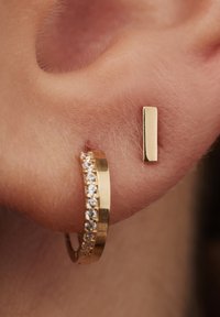Goldene Creolen mit einem Strassband neben einem schlanken, rechteckigen goldenen Steckerearring, vor einem Nahaufnahme-Hauthintergrund.