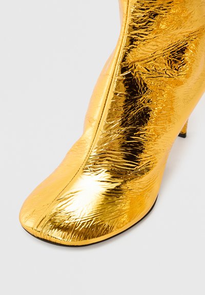 Χρυσό μποτάκι αστραγάλου με γυαλιστερή, τσαλακωμένη υφή, μυτερή μύτη και λεπτό τακούνι. Το υλικό αντανακλά το φως, δημιουργώντας μεταλλική εμφάνιση.