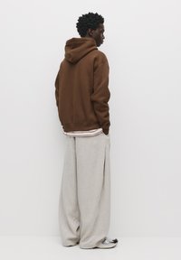 Brown hooded sweatshirt, loose beige trousers, and patterned sneakers. Minimal texture and design details visible from a side view.