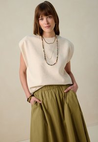 Top sans manches neutre avec épaulettes, associé à une jupe fluide vert olive. Trois colliers superposés et un bracelet accentuent le look.