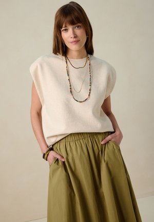 Neutrale mouwloze top met schouderkussens, gecombineerd met een vloeiende olijfgroene rok. Drie gelaagde kettingen en een armband accentueren de look.