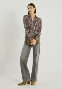 Blusa con estampado de leopardo y cuello, combinada con jeans grises de tiro alto y tacones en punta color coñac. Sosteniendo un pequeño bolso de mano marrón.