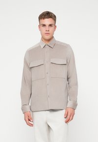 Camisa de botones color beige claro hecha de tela de pana con dos bolsillos delanteros, un cuello puntiagudo y mangas largas. Textura suave, patrón acanalado.