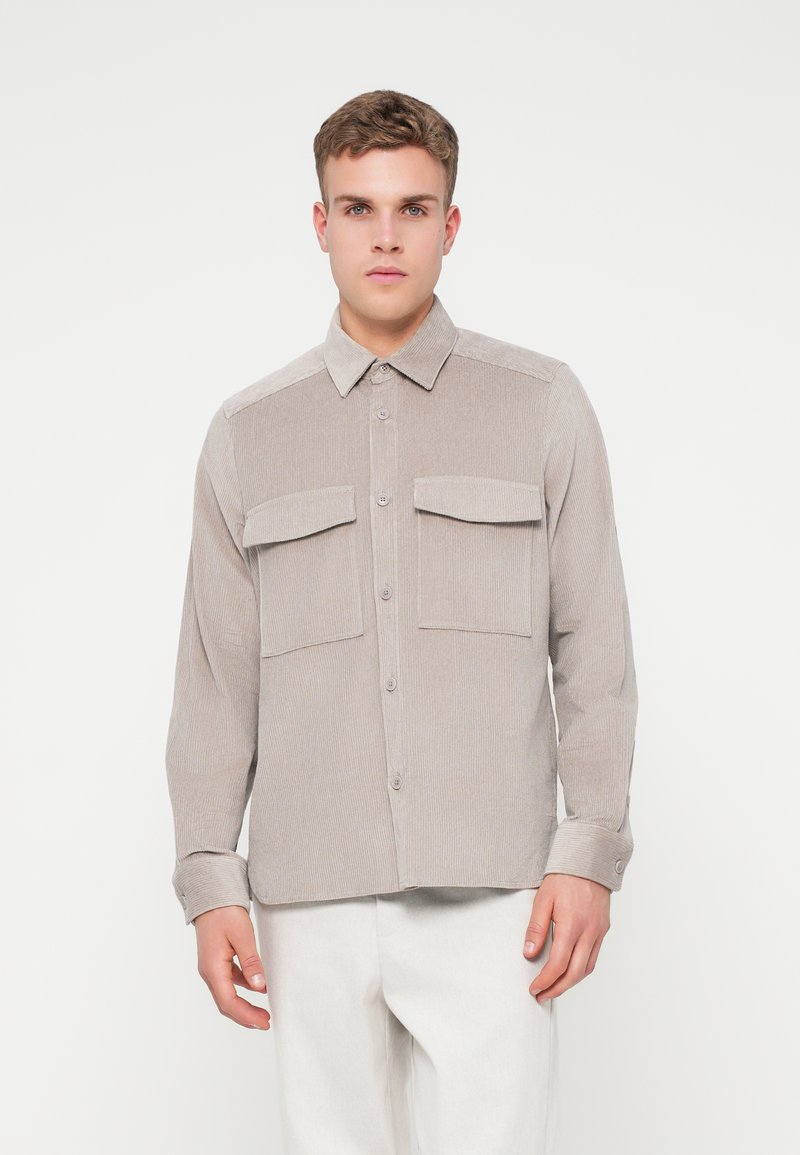 Camisa de botones color beige claro hecha de tela de pana con dos bolsillos delanteros, un cuello puntiagudo y mangas largas. Textura suave, patrón acanalado.