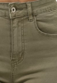 Pantalones de mezcla de algodón en verde oliva que cuentan con cierre de cremallera y botón, pasadores para cinturón y un bolsillo frontal con detalles de costura visibles.