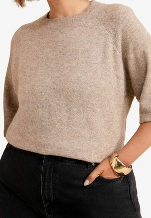 Vrouw draagt een beige gebreide trui met korte mouwen en zwarte high-waisted jeans met een gouden armband om haar pols, hand in de zak.