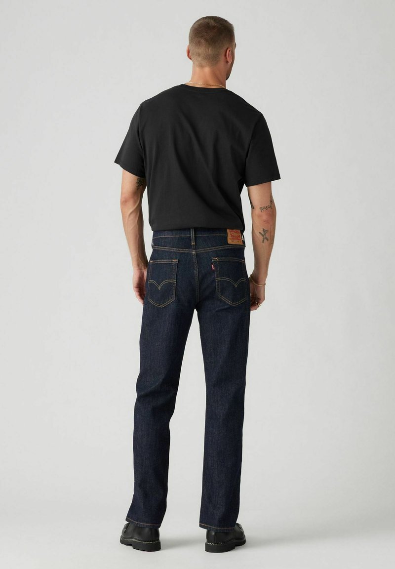 Jeans in denim blu scuro con taglio a gamba dritta, caratterizzati da una toppa in pelle sul retro e cuciture gialle a contrasto, indossati con una T-shirt nera.