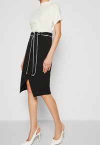 Černá tužková sukně s bílým páskem a asymetrickým lemem, spárovaná s krémovou blůzou s krátkým rukávem a bílými lodičkami na vysokém podpatku.