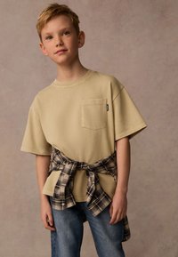 Beige t-shirt med kort ärm och framficka, bärs över blå jeans. En rutig skjorta knuten runt midjan ger kontrast med mörka toner.