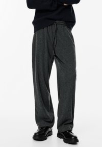 Személy laza, sötétszürke kötött nadrágot visel, amit zsinórral lehet összehúzni, fekete, vastag talpú cipővel és fekete pulóverrel, karba tett kézzel.